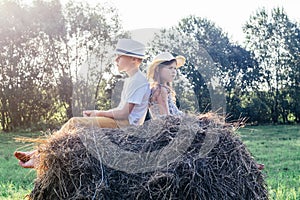 Portrait of two barefoot children boy and girl sitting their backs to each other on haystack in field. Kids wearing hats