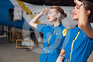 Portrait of two beautiful air stewardesses in stylish blue uniform smiling while looking up in the sky, standing
