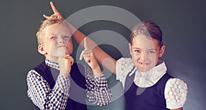 Portrait of two beautiful European kids boy and girl in school uniform standing next to the blackboard. Girl shows horns