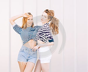 Portrait of two beautiful fashionable girlfriends in denim shorts and striped t-shirt posing nex to the glass wall. Girl holding