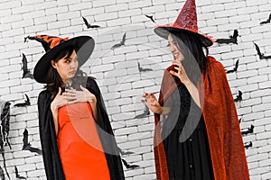 Portrait of two beautiful young asian woman in witch halloween costume on white brick background decorated with black paper bat