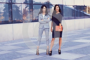 Portrait of two businesswomen dressed in a stylish formal clothes, standing in a downtown posing against a background of