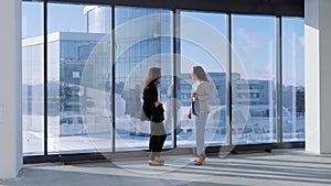 Portrait of two busineswoman in office standing in front of office buildings behind the window. Coworkers discussing