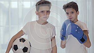 Portrait of two Caucasian twin brothers with black hair posing with ball and boxing gloves. Young schoolboys going in