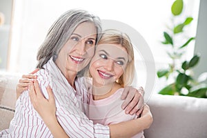 Portrait of two charming cheerful ladies sit on sofa hugging each other look far away beaming smile indoors