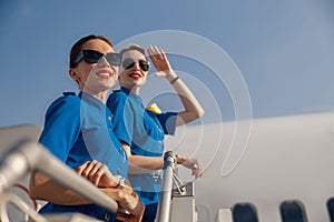 Portrait of two cheerful air stewardesses in blue uniform and sunglasses smiling away, standing together on airstair on