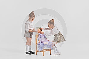Portrait of two cute little girls, children playing with doll, brushing hair isolated over grey background