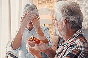 Portrait of two cute and old seniors at home having fun together. Mature man giving a gift at his wife for Christmas or