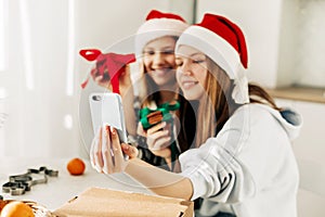 Portrait of two cute sisters in a Santa Claus Christmas hat in the kitchen, taking selfies with a smartphone, while