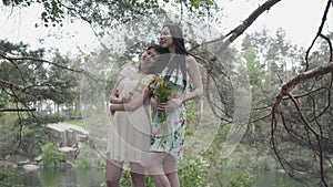 Portrait two cute young women in short summer dresses standing on rocky ground with wild flowers and looking forest