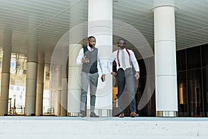 Portrait of two dark-skinned businessmen walking and talking in front of a modern building exterior. Friendly meeting