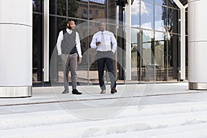 Portrait of two dark-skinned businessmen walking and talking in front of a modern building exterior. Friendly meeting