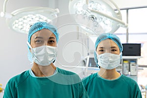 Portrait of two diverse female surgeons in caps and masks smiling in operating theatre