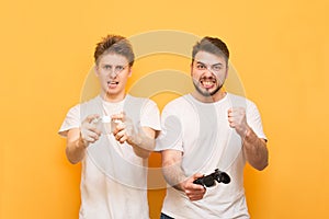 Portrait of two expressive gamers playing video games with joysticks in their hands on a yellow background, looking into the