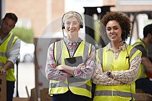 Portrait Of Two Female Workers Using Headsets In Distribution Warehouse With Digital Tablet