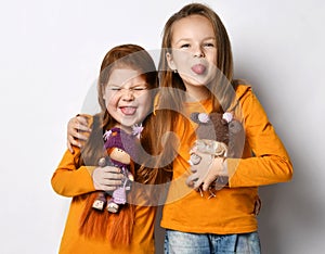Portrait of two frolic girls friends in orange shirts standing together with dolls hugging sticking out their tongues