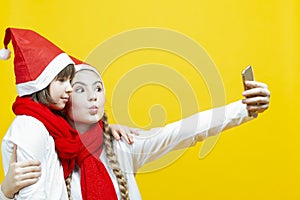 Portrait of Two Funny Caucasian Sisters Girfriends With Smartphone Taking Selfie Pictures in Santa Hat Over Yellow Background