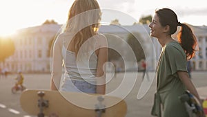 Portrait of two girls teenage friends with skateboard in sunshine in street european town laughing at something outside