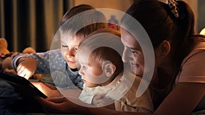 Portrait of two happy boys playing games on table computer with mother on bed at night. Family having time together, parenting,