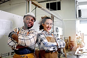 Portrait of two happy smiling carpenter man and woman wearing apron standing with arms crossed in woodworking furniture workshop,