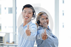 Portrait of two happy smiling diverse call centre telemarketing agents standing back to back and gesturing thumbs up for