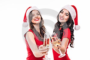 Portrait of two happy smiling girls in red dresses and santa hats holding glasses with champagne while toasting and looking at cam