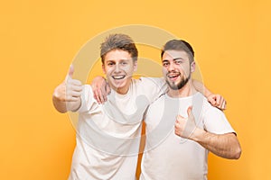 Portrait of a two happy young men showing thumbs up isolated over yellow background. Two happy friends in white t-shirts hugged by