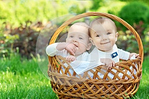 Portrait of two infant children in white suits sit in a basket on a picnic outdoor. Blond child is serious, boy with dark hair is