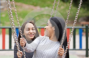 Portrait of two latin women friends are swinging on a swing set, smiling and laughing