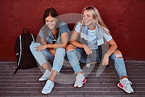 Portrait of a two laughing hipster girls sitting together on skateboard and leaning on a wall at sidewalk.