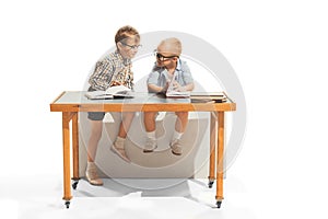 Portrait of two little boys, children, school pupils sitting at desk, studying, doing homework isolated over white