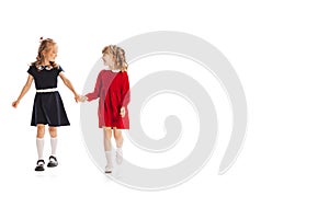 Portrait of two little girls, children in beautiful dresses holding hands, cheerfully walking isolated over white studio