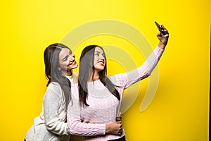 Portrait of two lovely girls dressed in sweaters standing and taking a selfie over yellow background