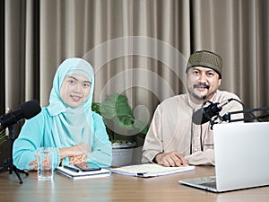 Portrait of two muslim radio hosts talking and smiling while sitting near microphones in broadcasting studio