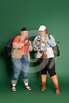 Portrait of two overweight women, friends in casual clothes with backpacks, eating hot dogs over green studio background