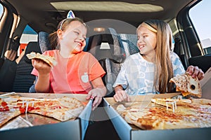 Portrait of two positive smiling sisters eating Huge just cooked italian pizzas sitting in child car seats on car back seat. Happy photo