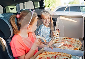 Portrait of two positive smiling sisters eating just cooked italian pizza sitting in child car seats on car back seat. Happy photo