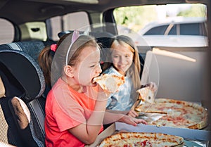 Portrait of two positive smiling sisters eating just cooked italian pizza sitting in child car seats on car back seat. Happy