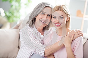 Portrait of two pretty cheerful ladies sit on sofa hugging hand on shoulder have good mood spend weekend indoors