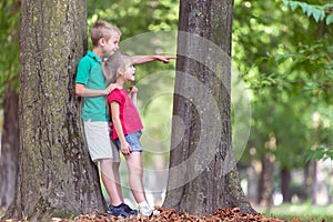 Portrait of two pretty cute children boy and girl standing near big tree trunk in summer park outdoors