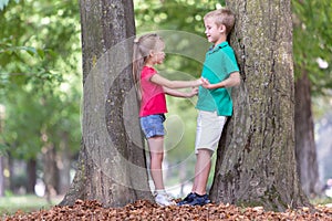 Portrait of two pretty cute children boy and girl standing near big tree trunk in summer park outdoors