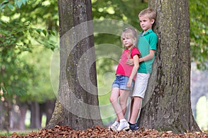 Portrait of two pretty cute children boy and girl standing near big tree trunk in summer park outdoors