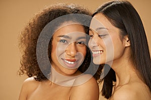 Portrait of two pretty mixed race young women with perfect smile posing together isolated over beige background