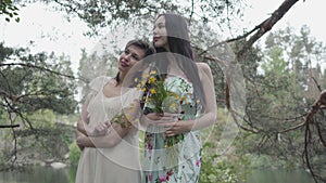 Portrait two pretty young women in summer dresses standing on rocky ground with wild flowers and looking forest enjoying