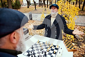 Portrait of two senior men playing chess in the park on a daytime in fall. Spreading hands. Win. Concept of leisure