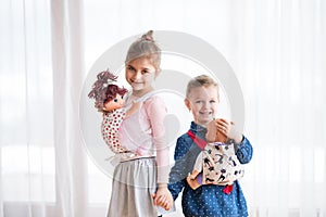 A portrait of two small girls standing and carrying dolls in baby carriers indoors.