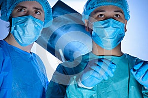 Portrait of two surgeons in sterile gowns, hats and masks. One surgeon, preparing to operate, holds a scalpel with a needle holder