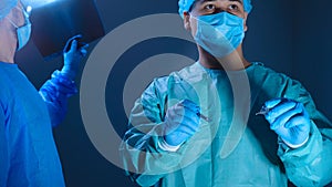 Portrait of two surgeons in sterile gowns, hats and masks. One surgeon, preparing to operate, holds a scalpel with a needle holder