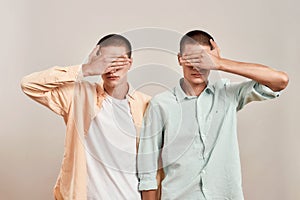 Portrait of two twin brothers covering eyes with hands while posing together isolated over beige background