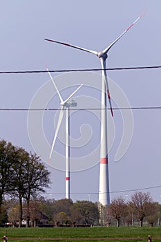 A portrait of two wind turbines standing in a field with red markings. In front of them there are some electricity power lines.